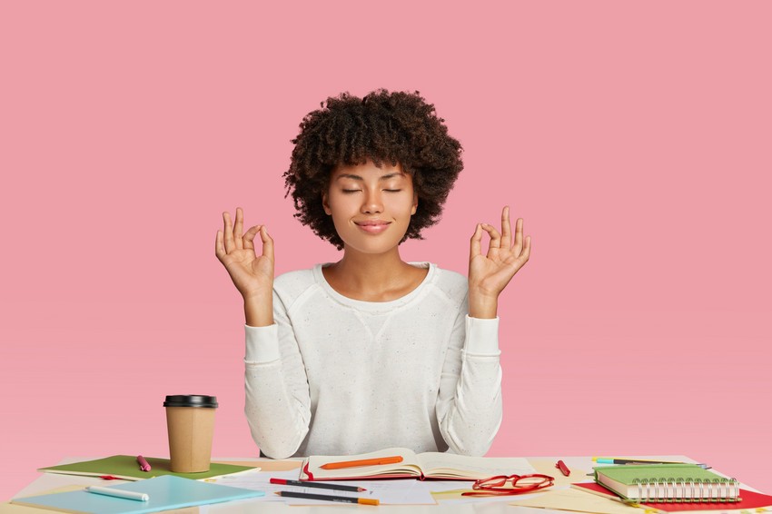 A person sitting at a table with notebooks, Ace QBank materials, and a coffee cup, meditating with eyes closed and fingers in a mudra position.