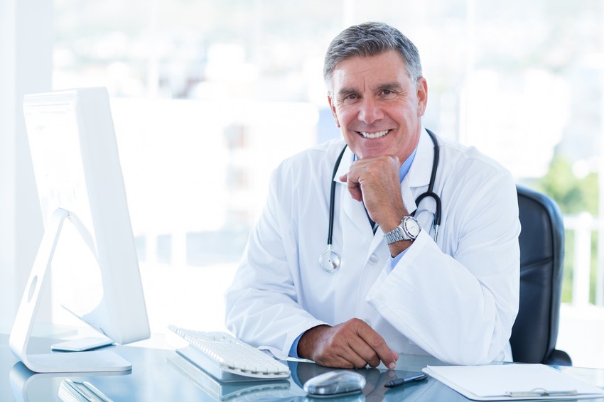 A smiling male doctor with a stethoscope around his neck is sitting at a desk with a computer, talking about MCCQE Part 1 exam preparation.