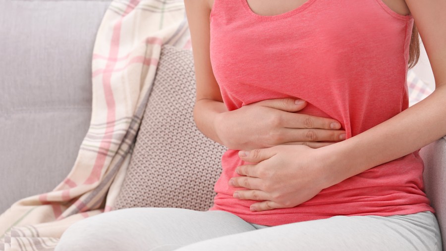 Woman in pink tank top clutching her abdomen in discomfort from cystitis, seated on a grey sofa with a cushion beside her.