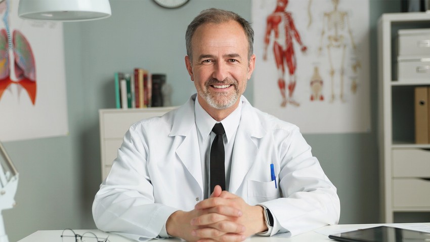 A smiling male doctor sitting at a desk in a medical office. Talking about how "Master MCCQE1 Ahead of The Curve" with anatomical charts and MCCQE1 exam preparation materials in the background.