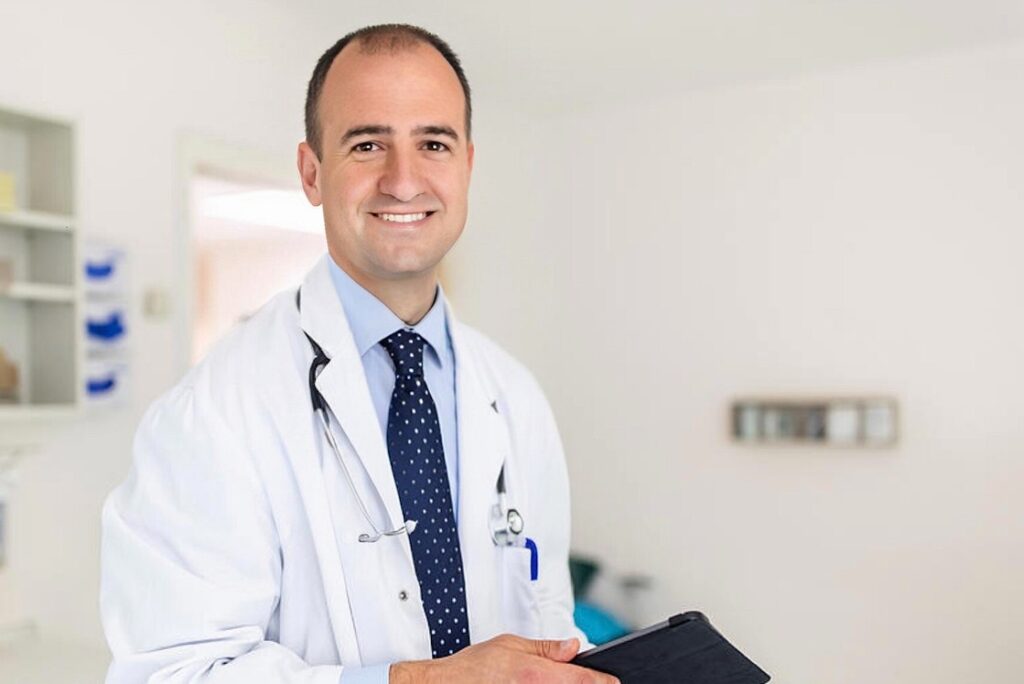 A smiling male doctor in a white coat and striped tie, holding a clipboard, standing in a bright hospital room, preparing for the MCCQE Part 1 and ready to deep dive into the best MCCQE1 Qbank.