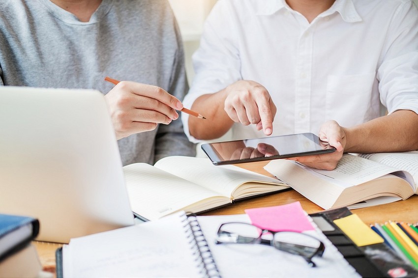 Two people collaborating at a desk with one holding a smartphone and the other pointing at its screen, surrounded by books and a laptop for Master MCCQE1 Ahead of The Curve.