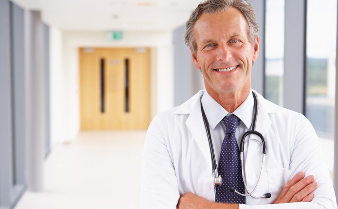 Smiling male doctor with crossed arms wearing a lab coat and stethoscope, standing in a hospital corridor, ready to talk about how to "Master MCCQE1 Ahead of The Curve."