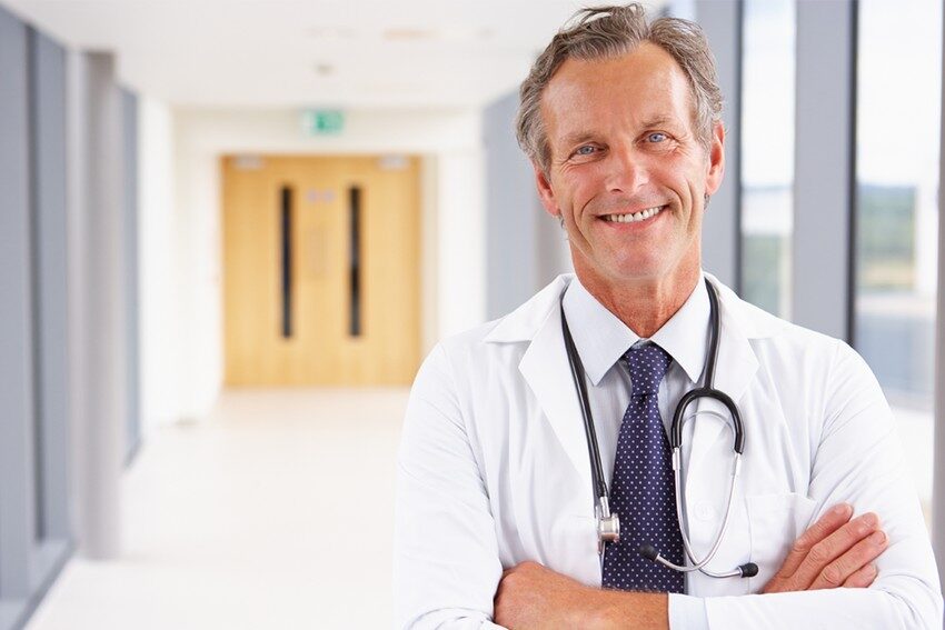 Smiling male doctor with crossed arms wearing a lab coat and stethoscope, standing in a hospital corridor, ready to talk about how to "Master MCCQE1 Ahead of The Curve."