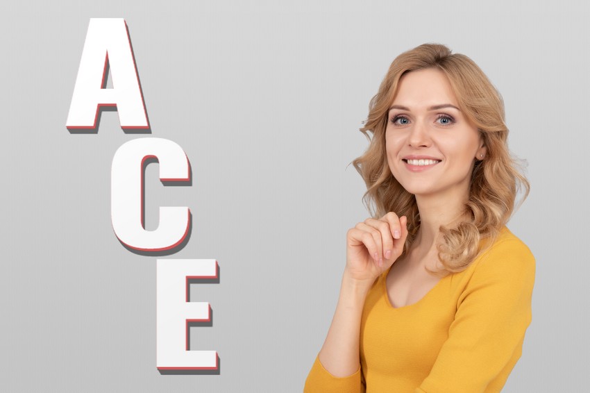 Smiling woman in a yellow top, contemplating floating 3d letters "ace" from Ace QBank beside her, against a grey background.