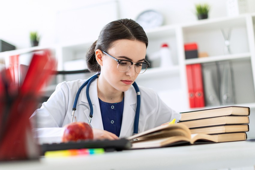 A medical student in a white coat and glasses, reading a book at her desk with a laptop and apple beside her for MCCQE1 exam preparation.