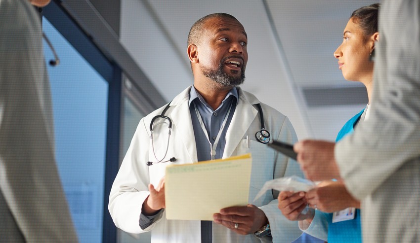 A middle-aged black male doctor with a stethoscope discussing with two colleagues in a hospital corridor, focusing on MCCQE part 1 Hidden cost and risk topics.