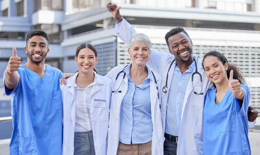 A diverse group of five smiling doctors in scrubs and lab coats, giving thumbs up outside a hospital saying "Master MCCQE1 Ahead of The Curve."