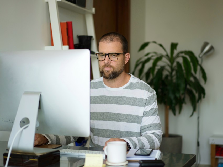 A man with glasses working intently at a desk with a computer, a cup of coffee, and a MCCQE Part 1 question bank.