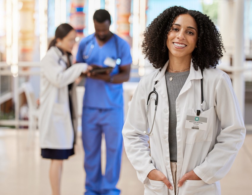 A confident female doctor in a white coat smiling at the camera, with a male and female colleague discussing MCCQE Part 1 question bank in the background in a hospital lobby.