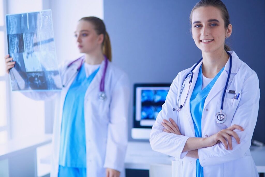 Two female doctors in a hospital, one examining an x-ray film and the other smiling at the camera, both wearing white coats and stethoscopes, discussing whether MCCQE part 1 is worth taking.