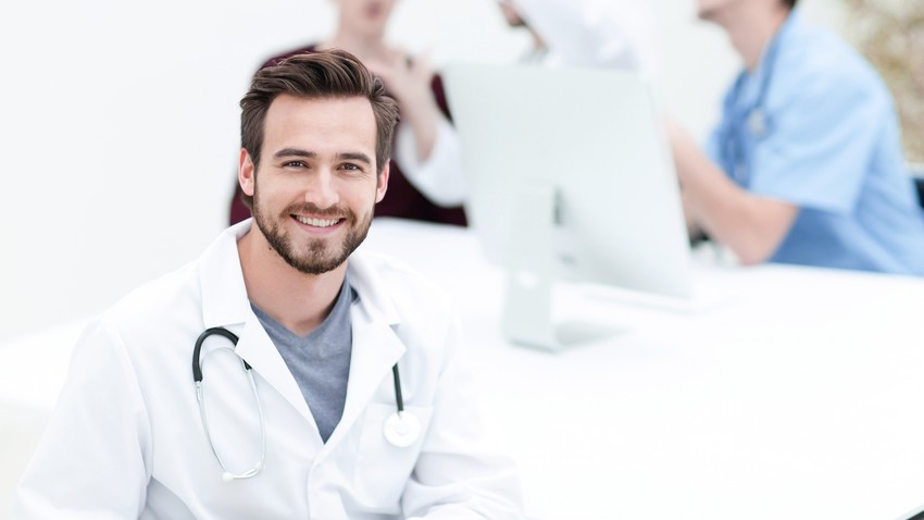 Smiling male doctor in a white coat with a stethoscope, sitting at a desk with medical staff and a computer in the background preparing for the MCCQE1.