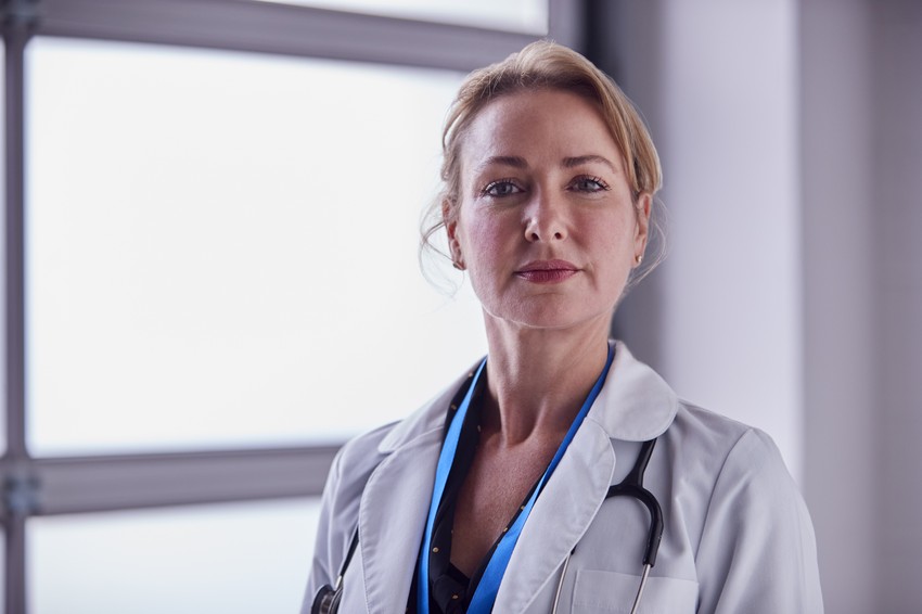 A confident female doctor with a stethoscope around her neck is standing in front of a window in a medical office, thinking about Ace QBank.