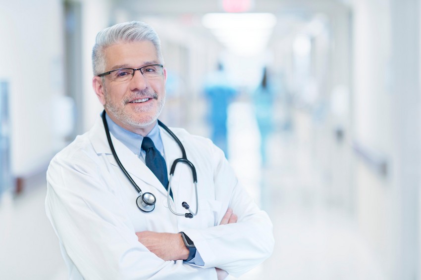 A male doctor with gray hair and glasses wearing a white coat and stethoscope standing confidently in a hospital corridor to talk about the MCCQE1