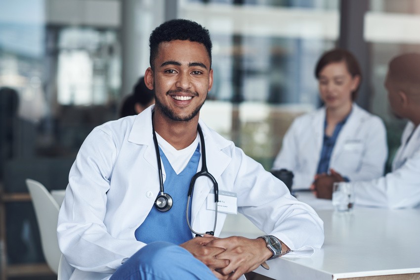 A smiling healthcare professional in a white coat and stethoscope sits at a table, possibly discussing Ace QBank strategies for MCCQE1 Prep, while two colleagues engage in conversation in the background.