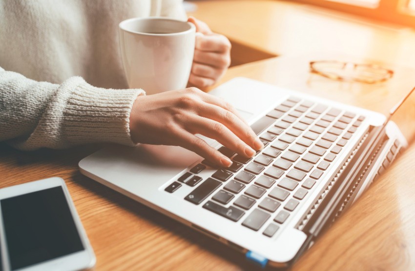 A person types on a laptop keyboard with one hand while holding a white mug in the other, next to a smartphone, Ace QBank notes, and a pair of glasses on a wooden desk. This Canadian question bank is perfect for MCCQE1 exam prep.