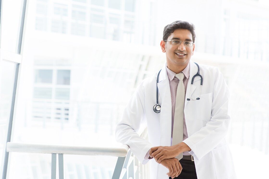 A doctor wearing a white coat and stethoscope stands indoors, leaning on a railing and smiling at the camera, confident after using Ace QBank for MCCQE1 Exam Prep.