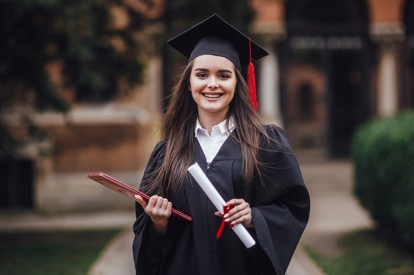 A young woman in a graduation cap and gown holds a diploma and a folder, smiling outdoors on a university campus, celebrating graduating college.