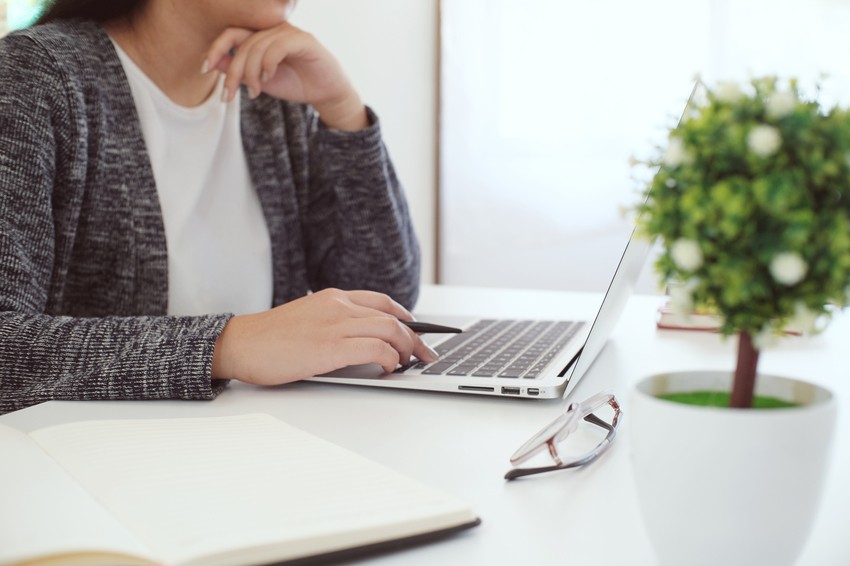 Person working at a desk with a laptop, notebook, eyeglasses, and a potted plant checks MCCQE1 Exam Dates on Ace QBank, resting their chin on one hand while typing with the other.