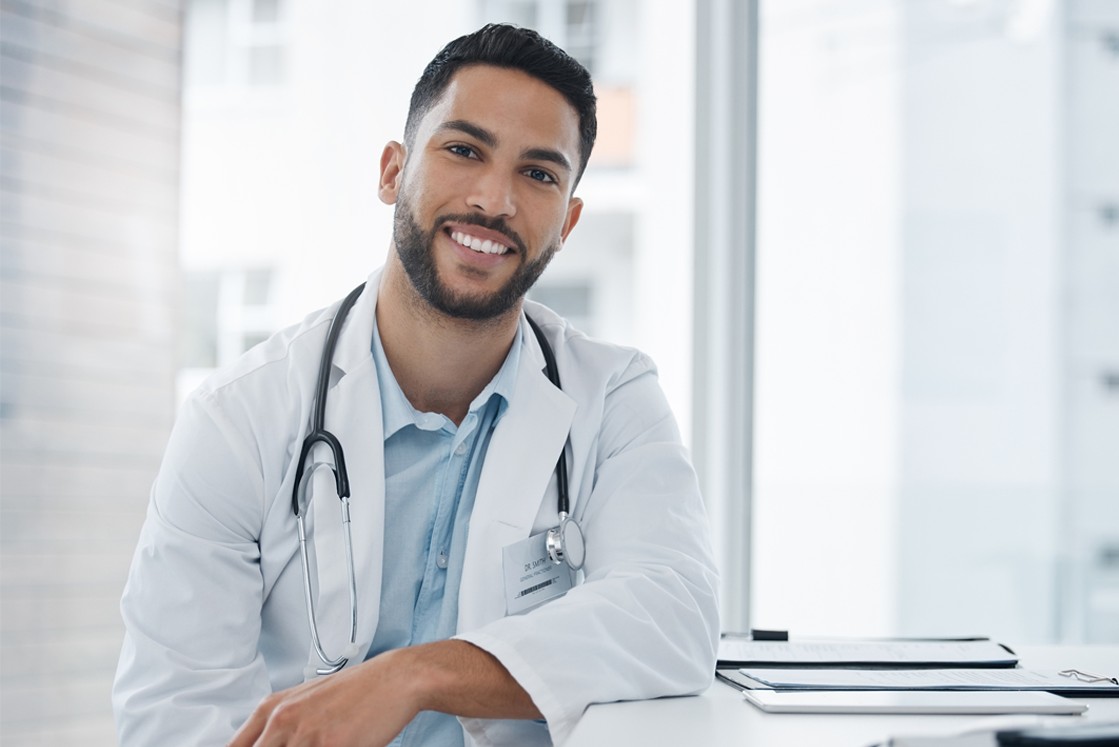 A male doctor wearing a white coat and stethoscope smiles while seated at a desk with an Ace QBank account open for the MCCQE1 exam prep and paperwork in a bright office.