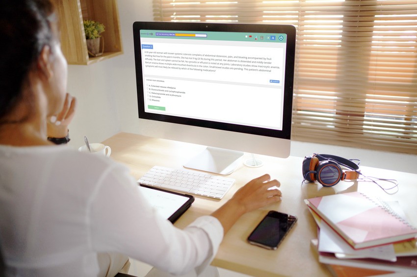 A person sits at a desk using a desktop computer, reading MCCQE1 Qbank text on the screen. Headphones, a notebook and a phone are also on the desk near the keyboard and mouse. Ready for more questions for better MCCQE1 exam prep.