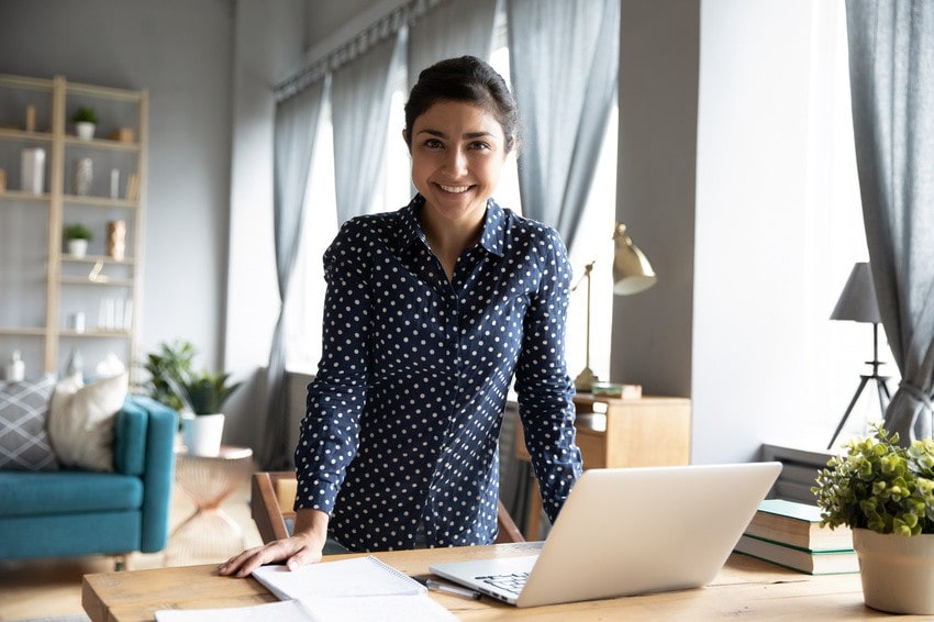 Woman in a blue polka dot shirt stands at a desk with a laptop, notebook, and papers in a bright, modern home office, focused on MCCQE1 exam preparation using an extensive Question Bank.