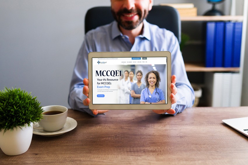 A man in business attire holds a tablet displaying an Ace QBank website for the MCCQE1 exam prep, with a cup of coffee and a plant on the desk in front of him.
