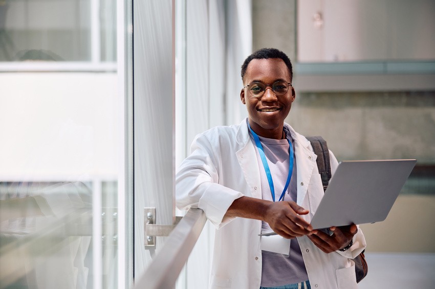 A person in a lab coat and glasses stands by a window holding a laptop, smiling, with a blue lanyard and backpack visible. Getting ready for MCCQE1 Exam with Ace QBank and inspired by the latest MCCQE1 Qbank New Update.