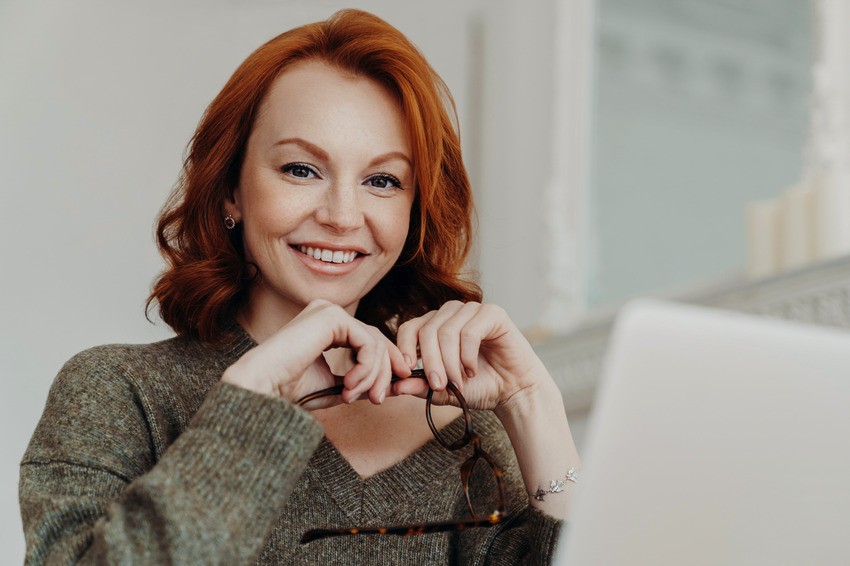A woman with red hair smiles at the camera while holding eyeglasses, sitting in front of a laptop. Ready to tackle More Questions for Better MCCQE1 Exam Prep with Ace QBank.