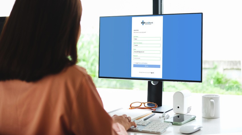 A person sits at a desk using a computer to fill out an online registration form, referencing Ace QBank for More Canadian Guidelines with New MCCQE1 Update. A notebook, pen, and glasses are on the desk.