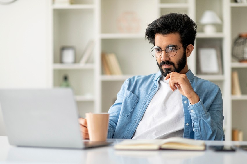 Man with glasses and a beard sits at a desk with a laptop, notebook, and mug, appearing thoughtful as he focuses on MCCQE1 Exam Preparation in a bright room with Ace QBank.