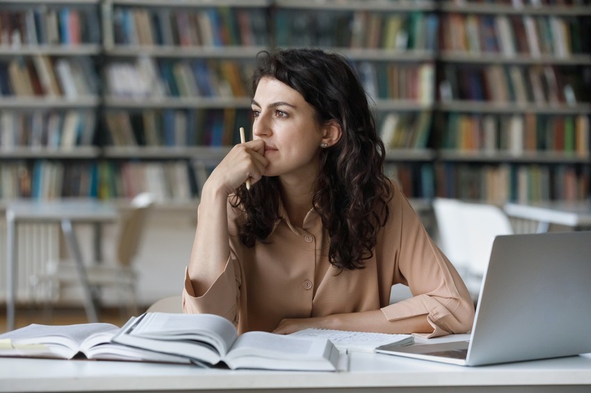 A woman sits at a desk in a library, surrounded by open books and a laptop, thoughtfully planning her MCCQE1 exam preparation with Ace QBank, a crucial step toward securing her medical licence in Canada.