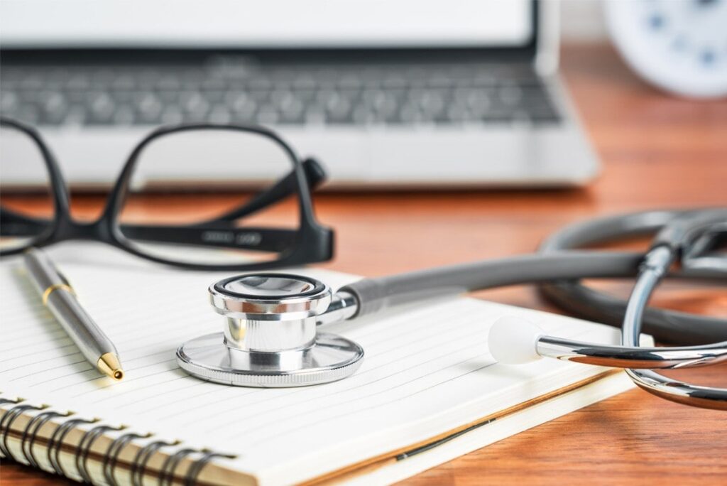 A stethoscope, notebook, pen, and eyeglasses rest on a wooden desk in front of an open laptop—perfect for your MCCQE1 exam preparation with Ace QBank and the last Chance to Secure Your MCCQE Part 1 Pass in 2025.