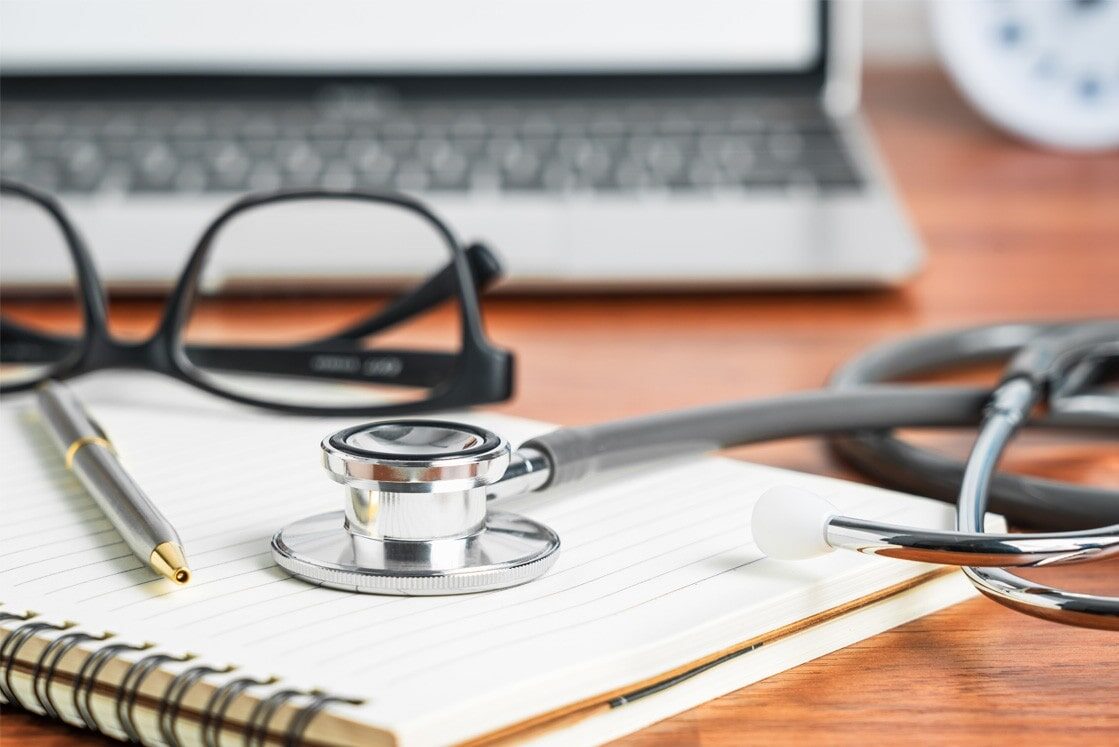 A stethoscope, notebook, pen, and eyeglasses rest on a wooden desk in front of an open laptop—perfect for your MCCQE1 exam preparation with Ace QBank and the last Chance to Secure Your MCCQE Part 1 Pass in 2025.
