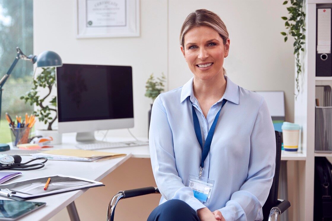 A woman with a name badge sits smiling at a desk in a modern office, studying MCCQE1 exam preparation materials on her computer. Making strides towards securing her medical licence in Canada, with documents, plants, and office supplies in the background.