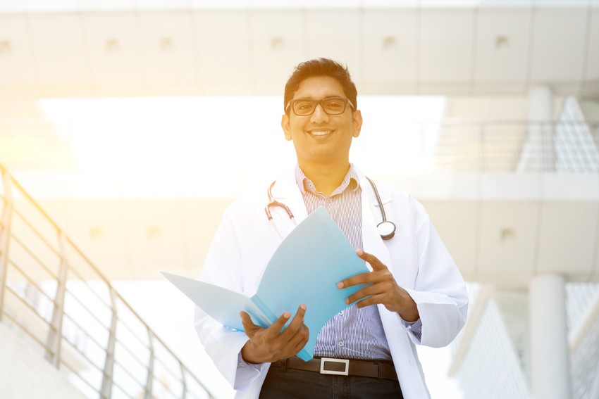A doctor wearing a white coat and stethoscope stands outdoors, holding a blue folder and smiling, with modern building architecture in the background. Ready for MCCQE1 exam preparation with Ace QBank.