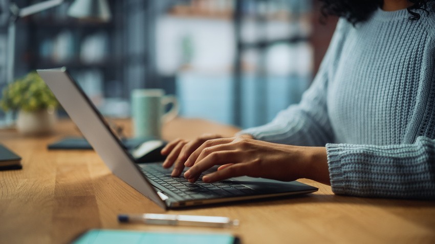 Person in a light blue sweater typing on a laptop at a wooden desk, with a pen and notepad nearby in a softly lit room. Focused on MCCQE1 exam preparation with Ace QBank to secure their medical licence in Canada.