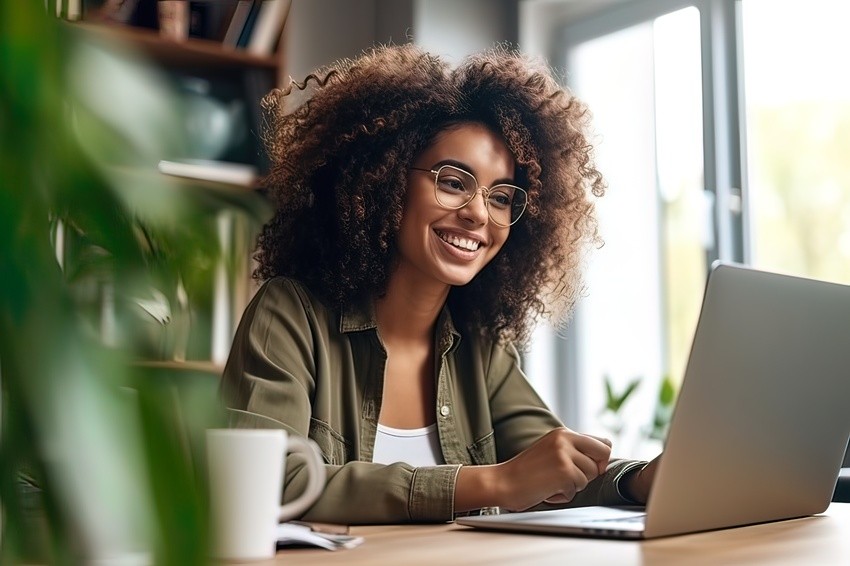A woman with curly hair and glasses sits at a desk, smiling while signing up for the Ace QBank demo for the MCCQE1 exam preparation on her laptop, with a mug and plants nearby.