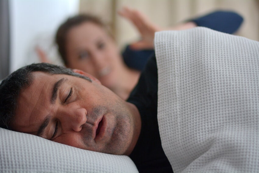 Man lying on his back in bed under a white textured blanket, mouth slightly open and head turned to the side, suggesting loud snoring or breathing obstruction; illustrative image for obstructive sleep apnea, Ace QBank clinical edge.