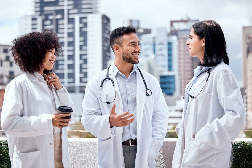 Three doctors in white coats stand outdoors in a city setting, talking and smiling. One, holding a coffee cup, shares tips about avoiding rookie mistakes that destroy MCCQE1 results while preparing for the MCCQE1 exam.