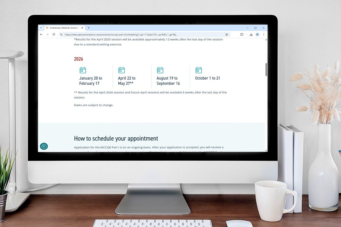 A computer monitor displays a scheduling webpage with 2026 MCCQE Part 1 Exam Dates and session details. A keyboard, mouse, and vase with dried flowers are on the white desk. Ideal for focused MCCQE1 Exam Preparation with Ace QBank.