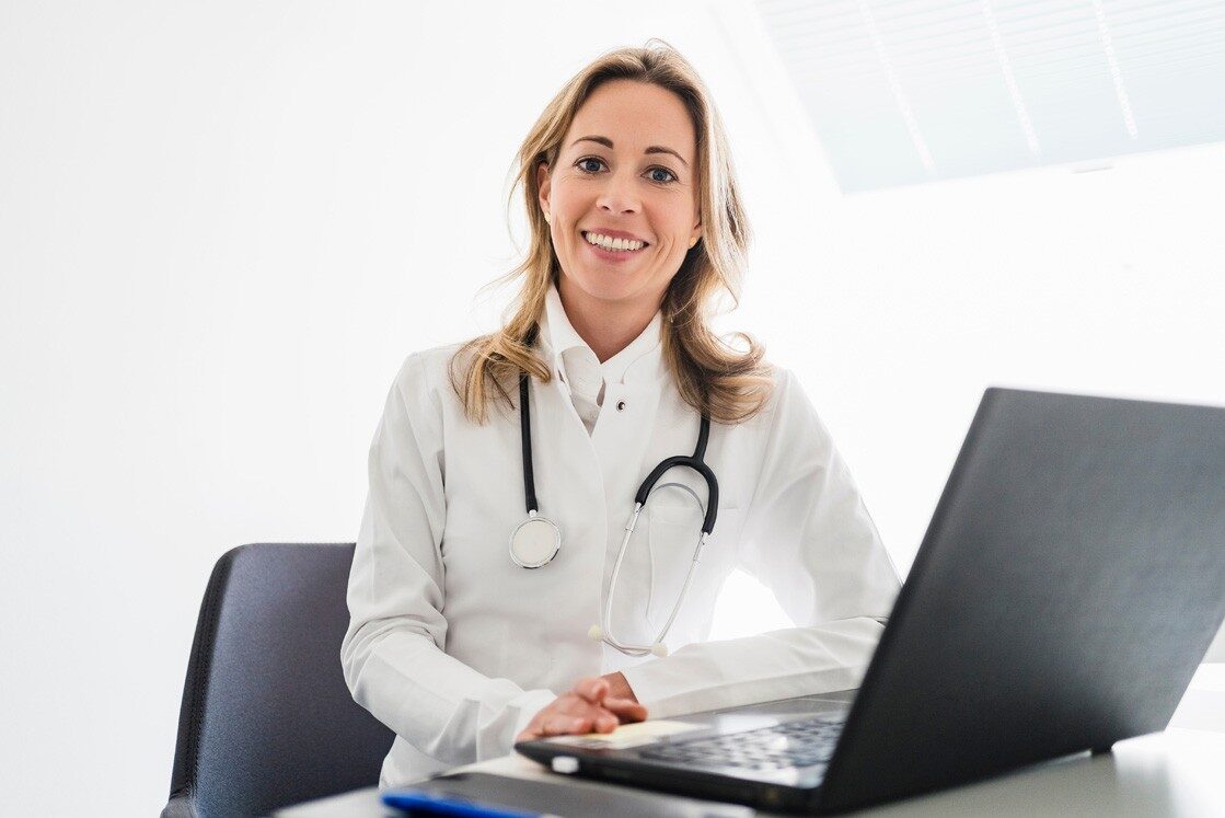 A doctor in a white coat with a stethoscope around her neck sits at a desk, smiling, with a laptop in front of her. Ready for MCCQE1 exam preparation using Ace QBank.