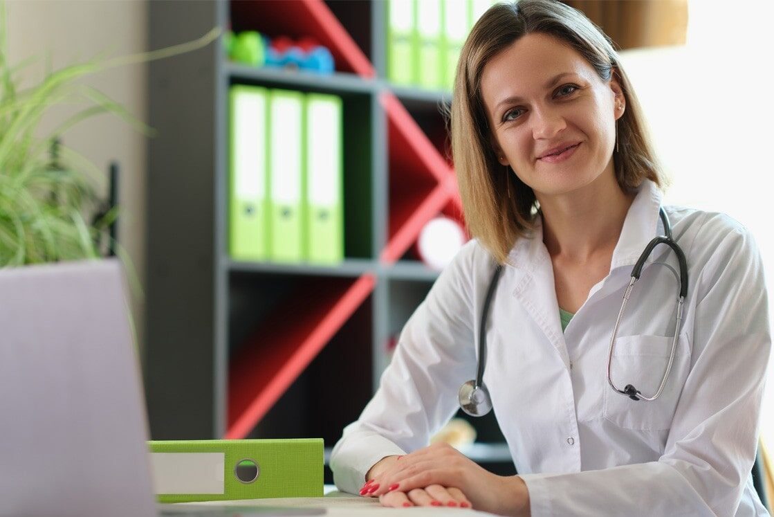 A woman in a white lab coat with a stethoscope sits at a desk, trying to explain how to avoid a rookie mistake that destroys MCCQE1 results while using Ace QBank to prepare for the MCCQE1 exam.
