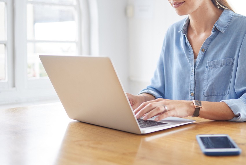 A person in a blue shirt types on a laptop at a wooden table with a smartphone nearby, reviewing Ace QBank to avoid the common mistake in the MCCQE1 Exam Prep.