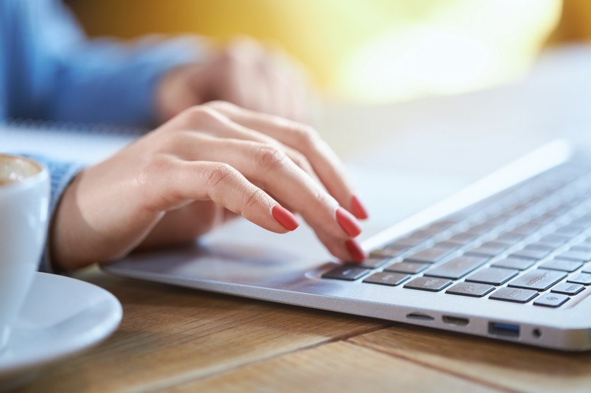 A person with red nail polish types on a laptop at a wooden table, preparing for the MCCQE1 exam with Ace QBank, while a coffee cup and notebook sit in the background.