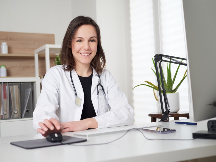 A woman in a white lab coat with a stethoscope sits at a desk, smiling at the camera. Behind her are a computer, files, and a potted plant. Perhaps she's just shared tips for avoiding the common mistake in MCCQE1 exam prep using Ace QBank.