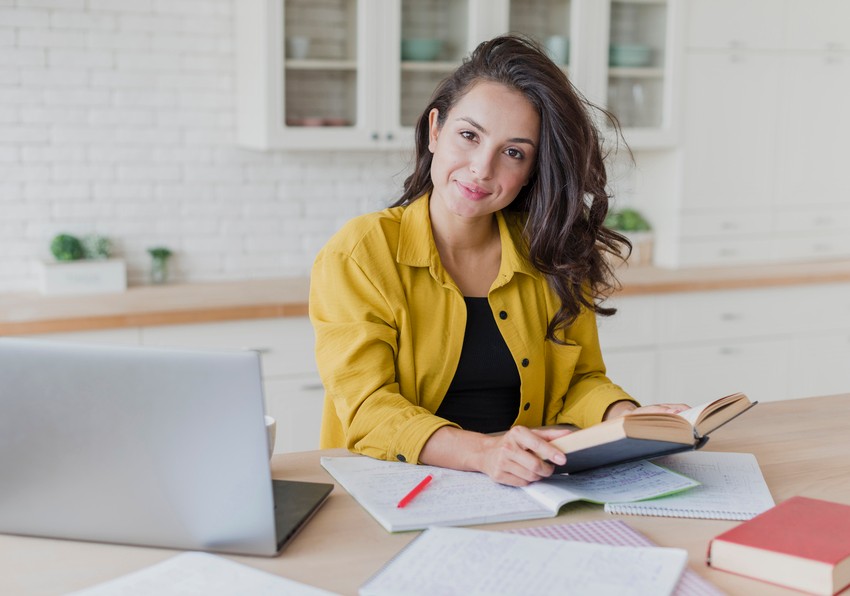 A woman sits at a table with an open book, notebook, and laptop, smiling at the camera in a bright kitchen while focusing on her MCCQE1 exam preparation, even after reading about the 2025 MCCQE Part 1 Exam Dates & Scheduling Gone.