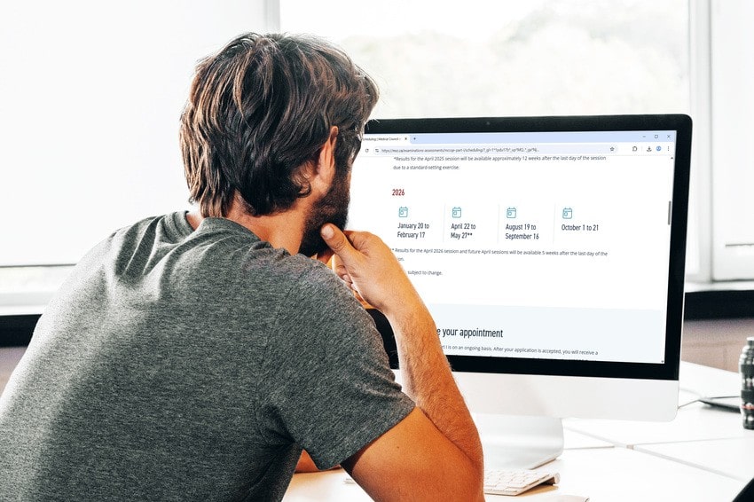 A man with short brown hair and a beard, wearing a grey t-shirt, sits at a desk looking at a large desktop monitor displaying a website with calendar dates for the 2026 MCCQE1 Exam Dates. he rests his chin on his hand and thinks about the Ace QBank for MCCQE1 exam preparation.
