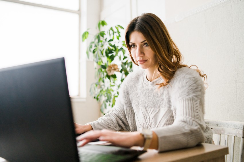 Woman with long brown hair wearing a light sweater sits at a desk, working on MCCQE1 exam preparation on her laptop in a bright room with a plant in the background.