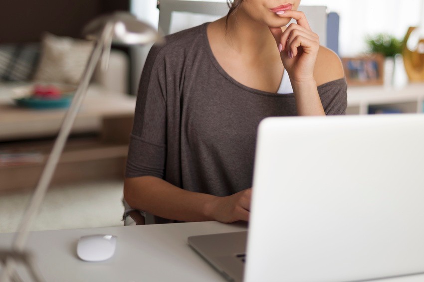 A woman sits at a desk with her hand on her chin, looking at a laptop screen as she reviews the 2026 MCCQE1 Exam Dates; a computer mouse and a lamp are on the desk.